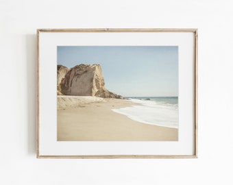 May include: A framed landscape photograph featuring a sandy beach with light beige sand, a large rocky cliff face on the left side, and gentle ocean waves washing onto the shore under a clear, pale blue sky. The frame is a light-toned wood.