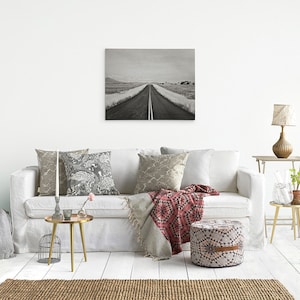 May include: A black and white photograph of a long, straight road leading to the horizon. The road is lined with trees and bushes, and the sky is cloudy. The photo is displayed on a white wall above a white couch with pillows and a throw blanket.