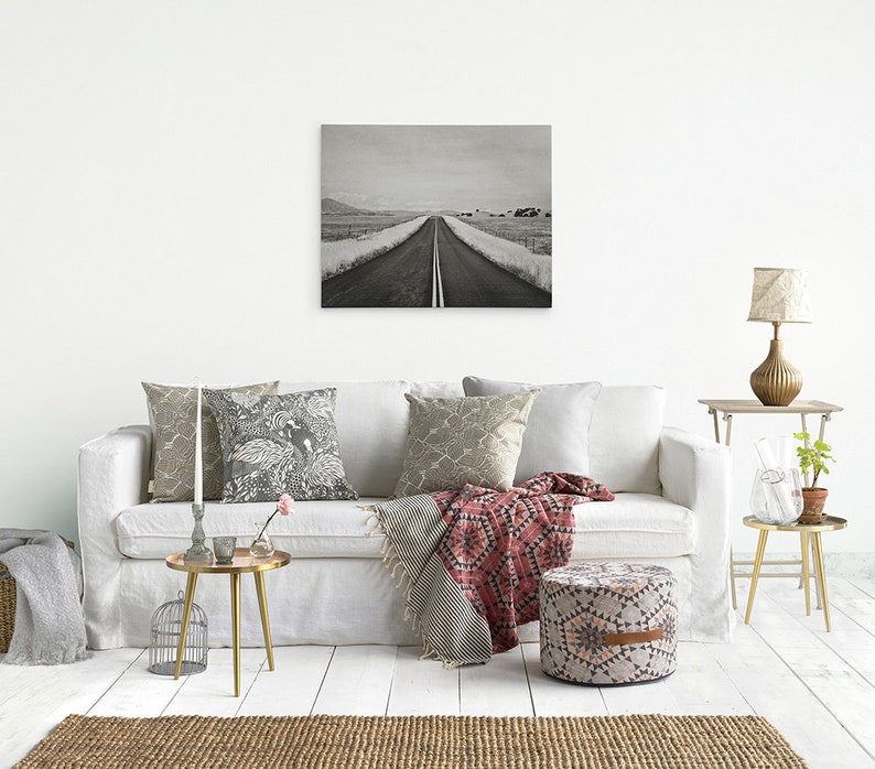 May include: A black and white photograph of a long, straight road leading to the horizon. The road is lined with trees and bushes, and the sky is cloudy. The photo is displayed on a white wall above a white couch with pillows and a throw blanket.