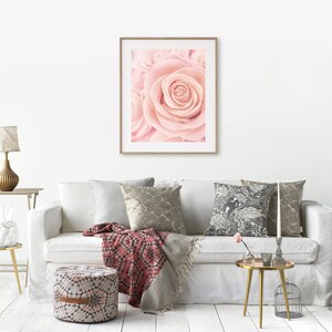 May include: A white sofa with a pink rose print framed artwork hanging above it. The sofa has a red and white throw blanket and a patterned pillow. There is a gold side table with a vase of flowers and a gold lamp on a table next to the sofa.