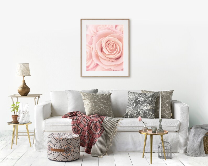 May include: A white sofa with a pink rose print framed artwork hanging above it. The sofa has a red and white throw blanket and a patterned pillow. There is a gold side table with a vase of flowers and a gold lamp on a table next to the sofa.