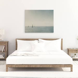 May include: A white bed with a wooden frame and white bedding. A framed photograph of a seascape with sailboats hangs above the bed. Two wooden nightstands with drawers are on either side of the bed.