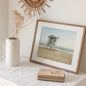 Puede incluir: Una fotograf&iacute;a enmarcada de una torre de salvavidas azul claro con el n&uacute;mero 38, situada en una playa de arena. La impresi&oacute;n est&aacute; en un marco de madera clara con paspart&uacute; blanco, junto a un jarr&oacute;n blanco y una pila de libros.