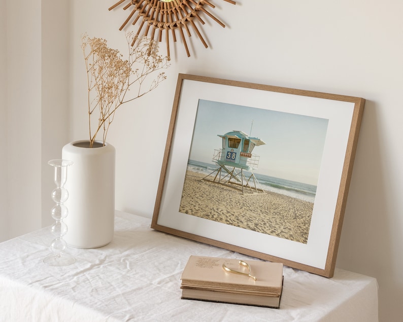 Puede incluir: Una fotograf&iacute;a enmarcada de una torre de salvavidas azul claro con el n&uacute;mero 38, situada en una playa de arena. La impresi&oacute;n est&aacute; en un marco de madera clara con paspart&uacute; blanco, junto a un jarr&oacute;n blanco y una pila de libros.