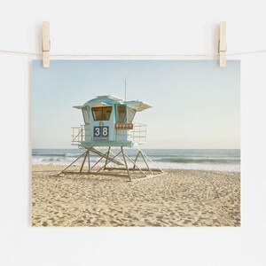 Puede incluir: Una torre de salvavidas de color azul claro se encuentra en una playa de arena con el oc&eacute;ano al fondo. La estructura presenta el n&uacute;mero 38 y el texto State Parks en su panel frontal, sostenida por un marco de metal sobre la arena.