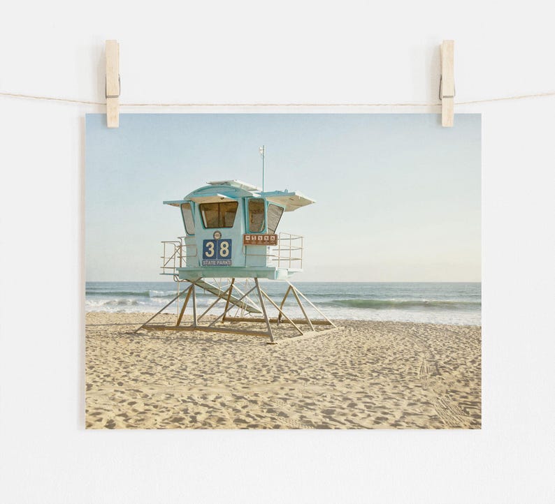 Puede incluir: Una torre de salvavidas de color azul claro se encuentra en una playa de arena con el oc&eacute;ano al fondo. La estructura presenta el n&uacute;mero 38 y el texto State Parks en su panel frontal, sostenida por un marco de metal sobre la arena.