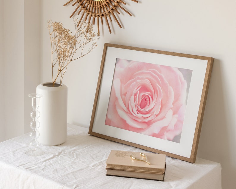 Pu&ograve; includere: Una stampa incorniciata di una singola rosa rosa con una cornice in legno marrone chiaro. La stampa &egrave; su un tavolo bianco con un vaso bianco e fiori secchi.