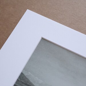 May include: A white mat board with a beveled edge and a rectangular opening for a photo or artwork. The mat board is on a brown surface.