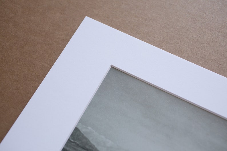 May include: A white mat board with a beveled edge and a rectangular opening for a photo or artwork. The mat board is on a brown surface.