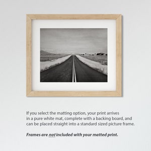 May include: A black and white photograph of a long, straight road leading into the distance. The road is lined with fields on either side, and the sky is overcast. The image is framed in a light wood frame.