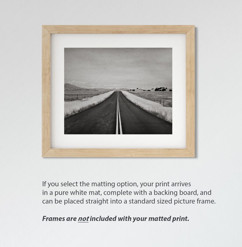 May include: A black and white photograph of a long, straight road leading into the distance. The road is lined with fields on either side, and the sky is overcast. The image is framed in a light wood frame.