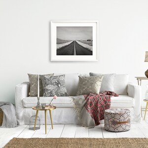 May include: A white sofa with a red and black throw blanket, a black and white patterned pillow, and a gray pillow. There is a gold side table with a glass vase and a plant. A gold round table with a candle holder and a small vase with a pink flower is in front of the sofa. A round, patterned ottoman is in front of the sofa. A black and white photo of a road is hanging on the wall above the sofa.