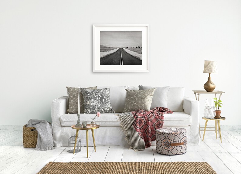 May include: A white sofa with a red and black throw blanket, a black and white patterned pillow, and a gray pillow. There is a gold side table with a glass vase and a plant. A gold round table with a candle holder and a small vase with a pink flower is in front of the sofa. A round, patterned ottoman is in front of the sofa. A black and white photo of a road is hanging on the wall above the sofa.