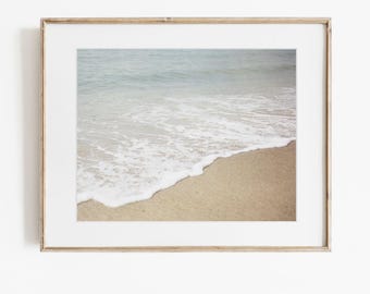 May include: A framed photograph of a shoreline featuring light tan sand meeting the edge of the ocean. White sea foam creates a curved line where the water washes onto the beach, with the calm, pale blue-grey sea extending into the distance.