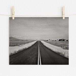 May include: A black and white photograph of a long, straight road stretching into the distance. The road is lined with tall grass on either side, and there are mountains in the background.