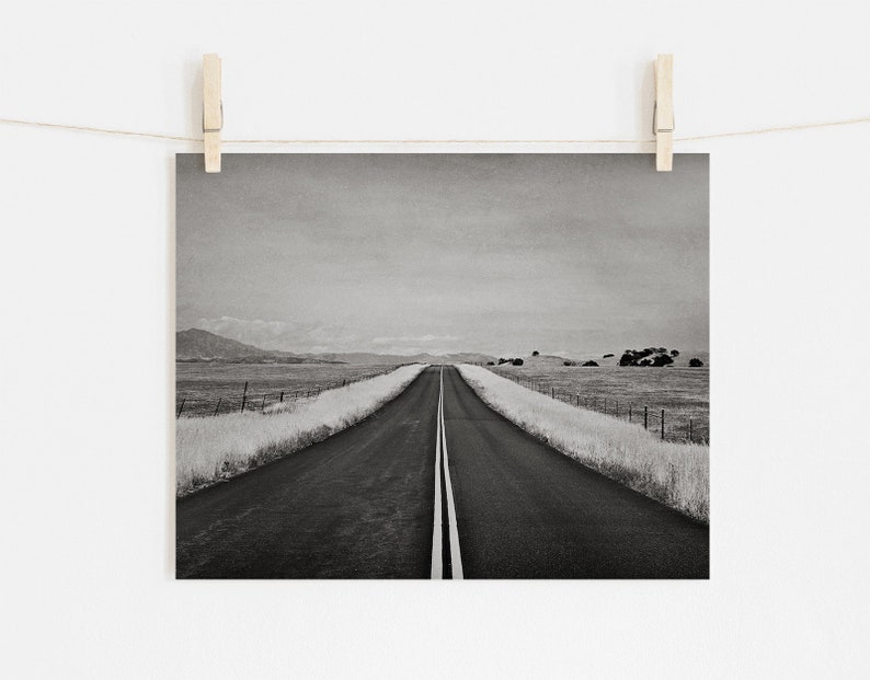 May include: A black and white photograph of a long, straight road stretching into the distance. The road is lined with tall grass on either side, and there are mountains in the background.
