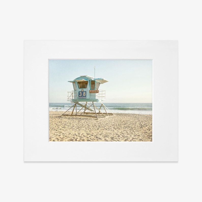 Puede incluir: Una impresi&oacute;n fotogr&aacute;fica montada que muestra una torre de salvavidas de color azul claro con el n&uacute;mero 38, situada en una playa de arena. La estructura se alza sobre pilotes met&aacute;licos frente a las olas bajo un cielo despejado.