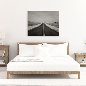 May include: A black and white photograph of a long, straight road disappearing into the distance. The road is lined with grass and trees on either side. The photo is framed and hanging on a white wall above a bed with white bedding.