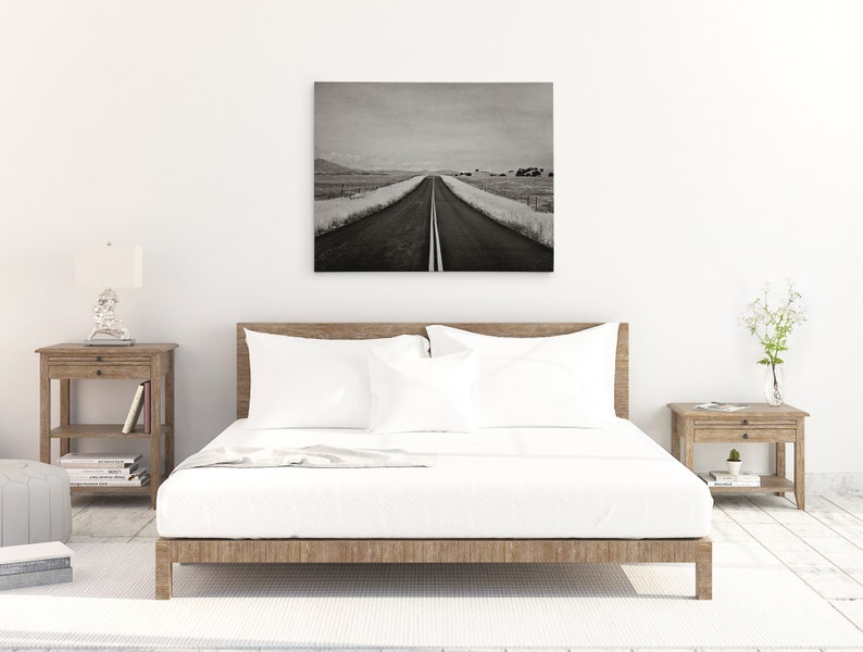 May include: A black and white photograph of a long, straight road disappearing into the distance. The road is lined with grass and trees on either side. The photo is framed and hanging on a white wall above a bed with white bedding.