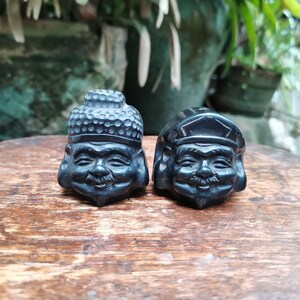May include: Two black wooden figurines of smiling faces with hats. The figurines are carved with intricate details and have a traditional Asian style.
