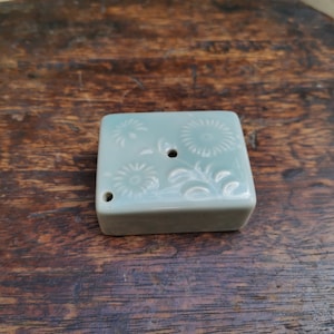 Könnte beinhalten: Ein kleiner, quadratischer, hellblauer Keramikkamin mit floralem Design und einem Loch in der Mitte.