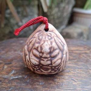 以下が含まれることがあります： しずく型の陶器製ベル。明るい茶色の仕上げで、濃い茶色の彫り込みにより様式化された顔が描かれています。ベルの上部には小さな穴があり、吊り下げ用の鮮やかな赤色の編み込みコードが通されています。