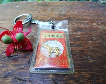 Op de afbeelding: Een gouden sleutelhanger met een klein, gedetailleerd ossenbedeltje. Het bedeltje is bevestigd aan een rode en gouden kaart met Japanse tekst die "金運牛開運" luidt. De sleutelhanger ligt op een houten oppervlak.