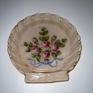 May include: A shell-shaped dish with a scalloped edge and gold trim. The dish features a hand-painted floral design of pink roses, green leaves, and a blue ribbon. The dish is likely made of porcelain or ceramic.