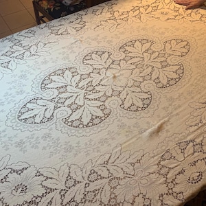 May include: Cream-colored, oval tablecloth with an intricate lace design. The lace features floral and leaf patterns, creating a delicate and elegant aesthetic. The tablecloth is spread across a table, showcasing its full design.