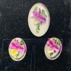 May include: A vintage jewelry set featuring an oval brooch and matching earrings. Each piece showcases a detailed orchid design with purple, yellow, and green accents encased in clear resin. The set is displayed on a black background.