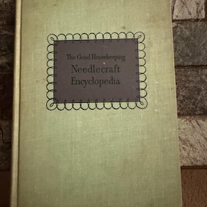 May include: A hardcover book titled The Good Housekeeping Needlecraft Encyclopedia with a light green fabric cover. The center features a dark gray rectangular label with a decorative scalloped border containing the book title in black text.