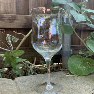 May include: A clear glass wine glass with a wide bowl and a long stem. The glass is empty and sitting on a gray concrete surface.