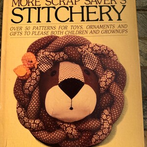 May include: A softcover book titled More Scrap Saver's Stitchery by Sandra Lounsbury Foose. The cover features a brown fabric lion head with a braided mane made of patterned brown fabrics and a small orange bird accent, with text detailing patterns.