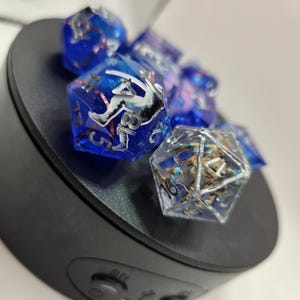 May include: A set of multi-faceted polyhedral dice made of translucent resin. The dice feature deep blue and clear colors with silver-colored numbering. One die contains a visible white and black graphic insert, while another displays internal brown textures.