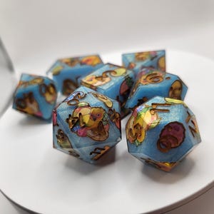 Könnte beinhalten: Ein Satz von sieben vielseitigen Würfeln mit hellblauer, durchscheinender Optik. Jeder Würfel zeigt farbenfrohe Illustrationen und goldfarbene Zahlen. Die Würfel liegen auf einer weißen Oberfläche, wobei einige scharf und andere unscharf sind.