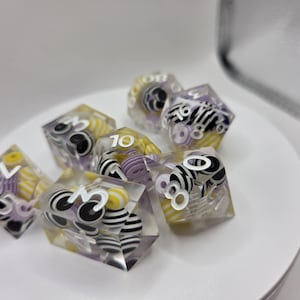 May include: A set of seven polyhedral dice made of clear resin, each containing small, colorful, circular beads in yellow, purple, and black-and-white striped patterns. The dice feature white engraved numbers on their faceted surfaces.