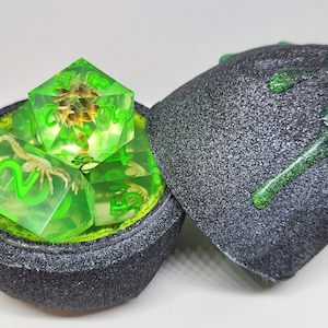 May include: A set of four green dice with a scorpion design inside. The dice are in a black and green container that looks like a rock.