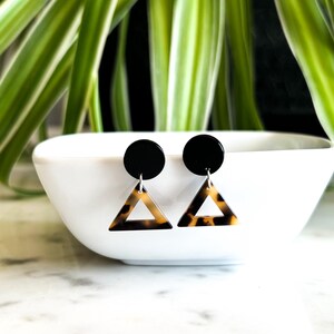 May include: A pair of black and brown geometric earrings with a triangle shape. The earrings have a black circle at the top and a brown and black triangle pendant.