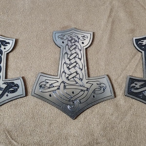 May include: Three silver-toned metal pendants shaped like Mjolnir hammers, each featuring an intricate, raised knotwork design with dark recessed detailing. The central pendant is larger than the two flanking it, and all display stylized animal motifs.