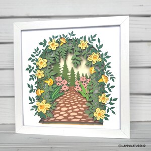 May include: A white framed paper art piece depicting a path through a forest with yellow flowers and green leaves. The path is made of brown stones and leads to a forest with green trees. The flowers are arranged in a circle around the path.