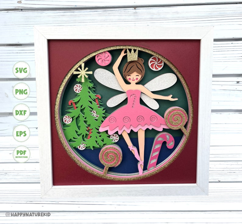 May include: A papercraft Christmas fairy with a pink tutu, wings, and a crown, surrounded by candy canes and a Christmas tree. The fairy is standing on a blue background with a gold glitter border.