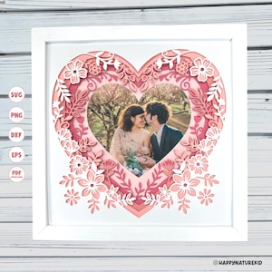May include: A framed paper art piece featuring a photo of a couple kissing, layered within a heart-shaped design. The heart is adorned with floral and leaf cutouts in shades of pink and white, set against a white frame.