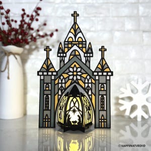 Puede incluir: Una linterna decorativa con forma de iglesia con un diseño de vidriera en gris, amarillo y negro. La linterna presenta una cruz en la parte superior y una escena de la natividad en el interior, iluminada por un brillo cálido. Un copo de nieve blanco y un jarrón con bayas rojas están en el fondo.