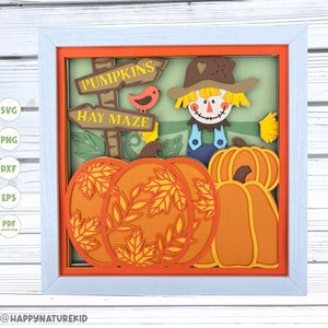 May include: A framed paper craft with a scarecrow, pumpkins, and signs that read "Pumpkins" and "Hay Maze". The scarecrow is wearing a brown hat, blue overalls, and a straw beard. The pumpkins are orange and have leaves on them.