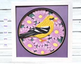 Puede incluir: Un pájaro amarillo y negro con una franja blanca en el ala está posado en un círculo de flores moradas con centros amarillos. Las flores están recortadas en papel y superpuestas para crear un efecto 3D. La imagen está enmarcada en un marco blanco.