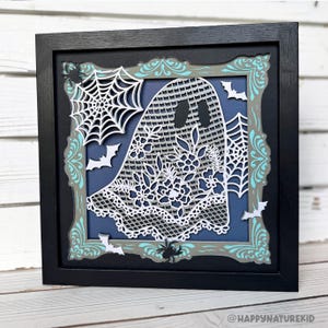 May include: A framed Halloween-themed paper art piece. The artwork features a white ghost with floral and lace details, surrounded by spiderwebs and bats. The frame is black with a decorative gray and teal border.