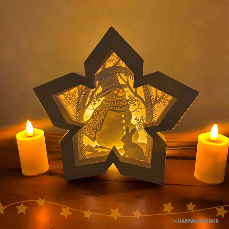 May include: A white wooden star-shaped light-up decoration with a cut-out design of a snowman and a rabbit in a winter scene. The star is illuminated from behind by a warm light source, creating a glowing effect.