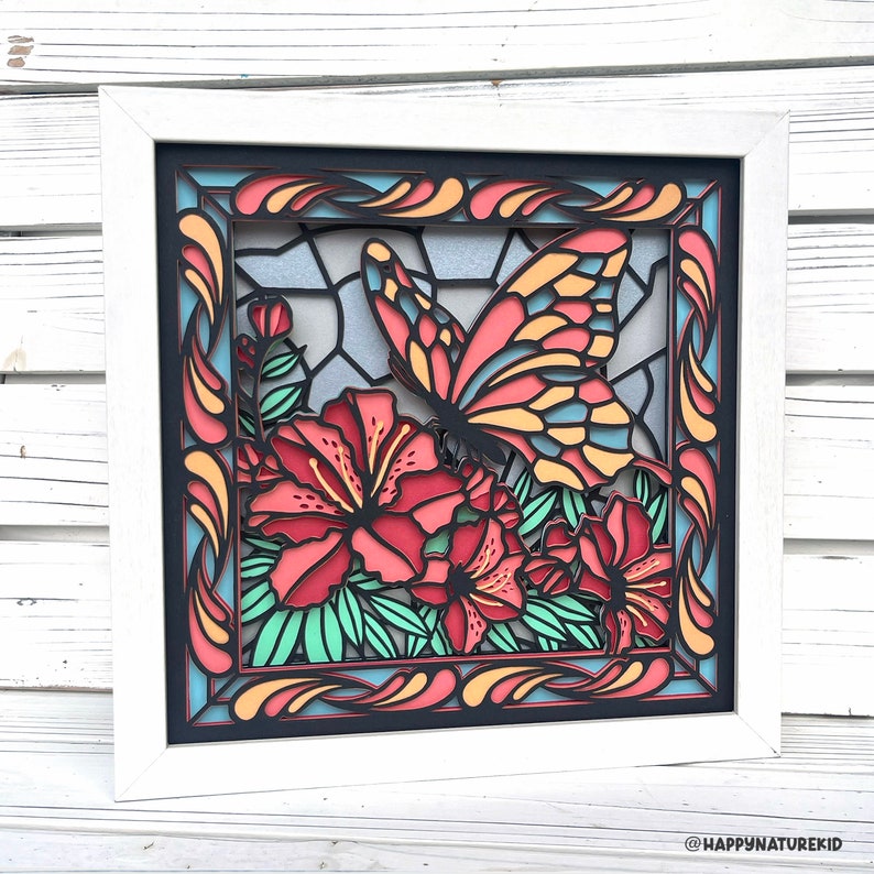 May include: A white framed wall art piece with a black background featuring a colorful butterfly and flowers. The butterfly is red, yellow, and blue with black outlines. The flowers are red with green leaves. The frame has a decorative border with swirls and lines in orange, yellow, and blue.