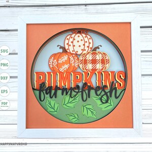 May include: A white frame with a layered paper craft design featuring three pumpkins with different patterns. The pumpkins are stacked on top of each other and surrounded by green leaves. The text "Pumpkins farm fresh" is written in black letters below the pumpkins.