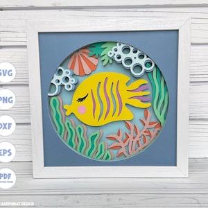 May include: A white frame with a blue background and a yellow fish with purple stripes and pink cheeks. The fish is surrounded by green and pink seaweed and coral.